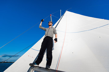 Positive skilled young male sailor with beard wearing sunglasses adjusting sail on boat and examining it while fixing sail on boom.