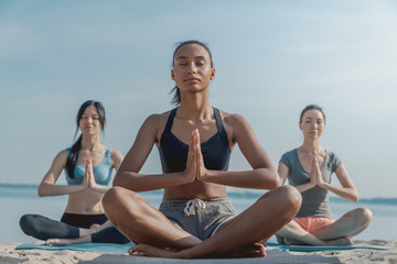 Three women in fitness wear doing yoga and meditation sitting at the seashore.