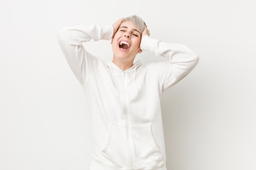 Young curvy woman wearing a white hoodie laughs joyfully keeping hands on head. Happiness concept.