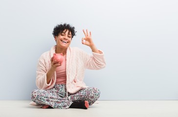Young african american woman sitting with a piggy bank cheerful and confident showing ok gesture.
