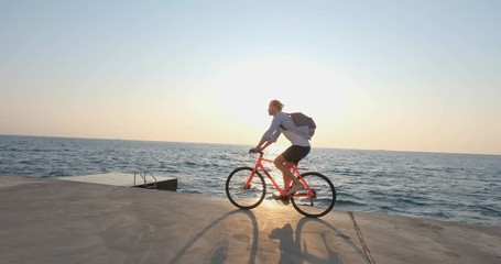 Young handsome male in casual wear ride on the colorful bicycle on the morning beach against beautiful sunset and the sea - Powered by Adobe