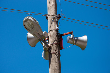 Loudspeakers and a street lamp on a pole with electric wires against a blue sky.
