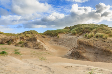 Dune valleys with deep wind holes carved out by heavy storm with swaying marram grasses with scattered clouds against blue sky