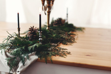 Fir branches with pine cones and vintage candlestick with burning black candles on wooden table. Stylish rustic christmas arrangement for festive dinner. Christmas rural table decor for feast