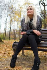 2017.10.21, Moscow, Russia. A young attractive woman wearing fur black jacket holding book sitting on the bench in falling park. Walking around autumn park.