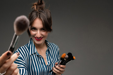 Portrait of young smiling woman in striped blouse with makeup brushes in her hands and showing one...