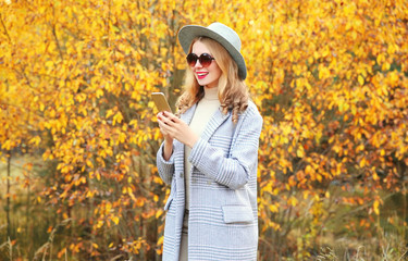 Autumn portrait stylish happy smiling woman holding phone wearing gray coat, round hat on yellow leaves background