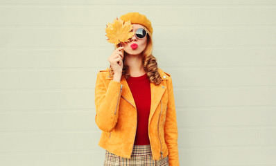 Autumn portrait woman holding in her hands yellow maple leaves hiding her eye, girl blowing red lips sending sweet air kiss over gray wall background