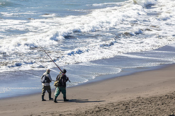 Two traditional fishermen going back home after some fishing at Matanzas beach in the Chilean coastline. The waves smash the sandy beach while they walk after a working day at the outdoors
