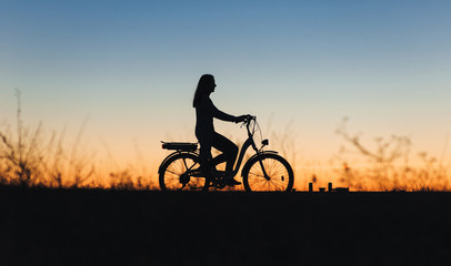 Silhouette of a young girl on the e-bike or electric bicycle on the sunset background. Country...
