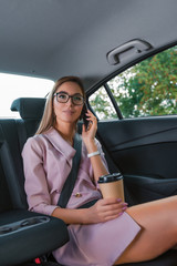 Fototapeta na wymiar Beautiful business lady woman rides passenger compartment passenger car VIP taxi, fastens smartphone her hands, rings, listens road, important call from secretary. Long hair, cup coffee tea drink.