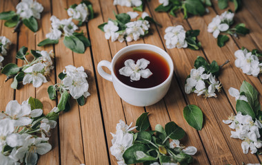 White cup of tea with petals of an apple tree on a wooden background, view from above.