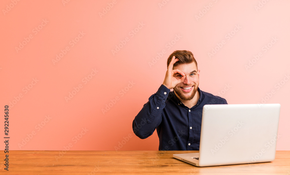 Wall mural Young man working with his laptop excited keeping ok gesture on eye.