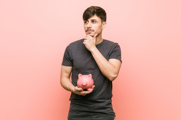 Young hispanic man holding piggy bank looking sideways with doubtful and skeptical expression.