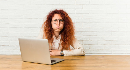 Young redhead curly woman working with her laptop blows cheeks, has tired expression. Facial...