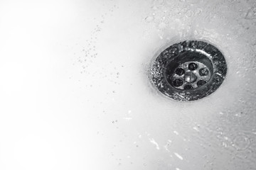 A stream of clean water flows into the drain stainless steel sink in white tones. Close up of drain and water in bathroom