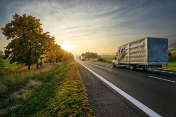Delivery van and truck driving on the asphalt road towards sunset around line of trees in misty...