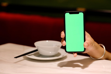 One hand showing green screen smartphone in Chinese restaurant. Blur chopsticks, dish and bowl background