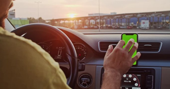 Close-up young man using smartphone mock-up greenscreen on dashboard driving a car in slow motion at sunset.