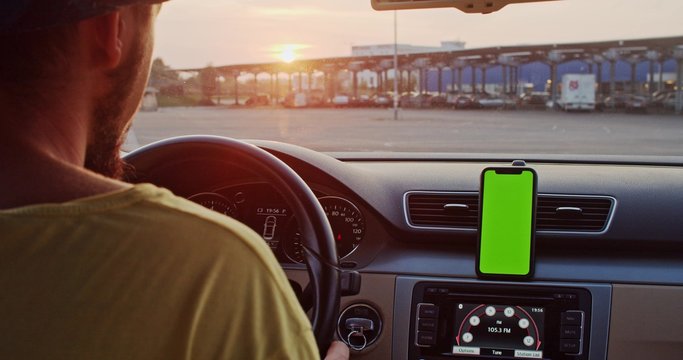 Close-up Young Man Inside Car Using Mounted Smartphone Navigator On Greenscreen Mock-up Display Driving At Sunset In The City.