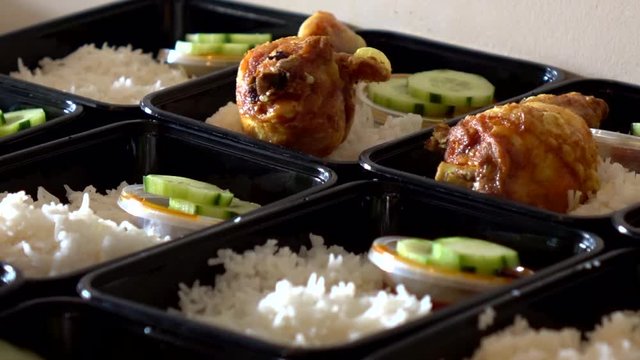 A row of Nasi Lemak in lunch boxes container while hands put a hot crispy chicken roasted on it.