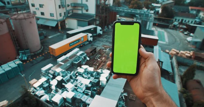Close-up of male hand holding vertical smartphone greenscreen mock-up technology on industrial background.