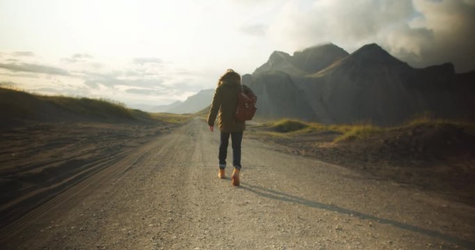 Male hiker with backpack walks dusty road in gloaming light at northern countryside