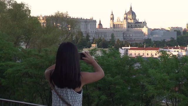 Locked off view of Japanese girl taking pictures of the Royal Palace of Madrid in the distance with smartphone CROP