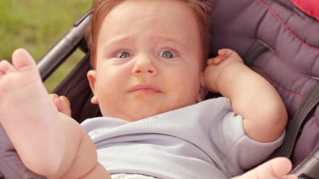 White unhappy infant with dark hair and dirty face is sitting in stroller on fresh air. Mother takes it out of the stroller