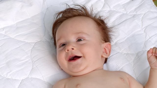 White smiling little child with dark thick hair and dark eyes is lying topless on the blanket on open air and observes the area. It is moving head and fingers. It raises left and right hand