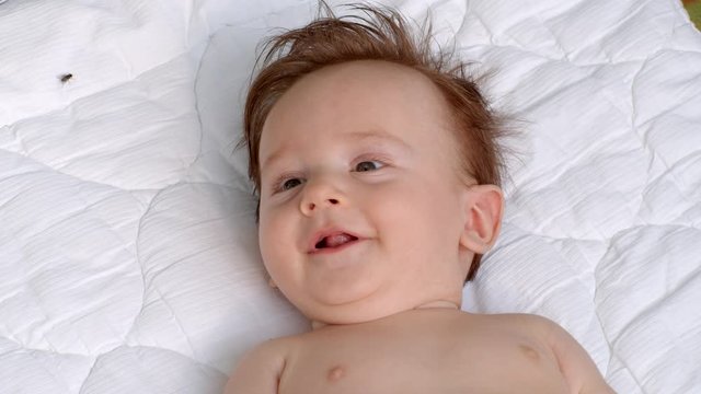 White smiling little child with dark thick hair and dark eyes is lying topless on the blanket on open air and observes the area. It is moving head and fingers. It raises left and right hand