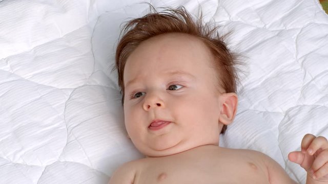 White smiling little child with dark thick hair and dark eyes is lying topless on the blanket on open air and observes the area. It is moving head and fingers. It raises left and right hand