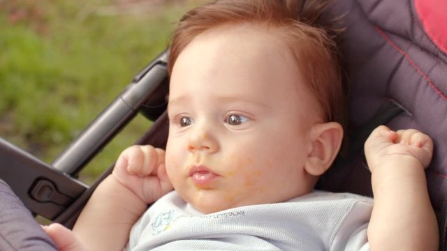 White excited infant with dark hair and dirty face is sitting in stroller on fresh air. It is observing area and moving its arms and legs