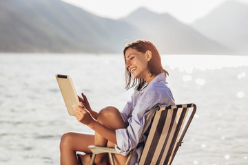 Smiling woman sitting on deck chair by the sea using tablet on a sunny day