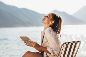 Smiling woman sitting on deck chair by the sea using tablet and credit card on a sunny day