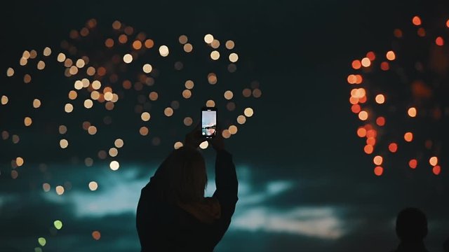 Happy woman shoots beautiful salute in the night sky of a megalopolis on a smartphone. Silhouette of people watching explosive and colorful fireworks in the evening sky. Holiday event and celebration.