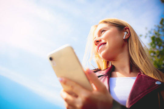 Young beautiful smiling adult Caucasian girl walking outdoor in the park having chat communication with wireless headphones mobile reading talking listening using with her hand on sunset at summer