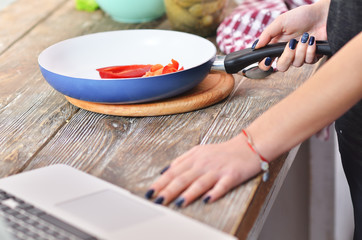 A young girl in a black t-shirt with laptop stirs pepper in the pan on a light wooden board on a gray table in a kitchen.