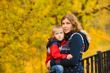 Fototapeta na wymiar Mom with her son in warm clothes, on bridge in an autumn park with yellow leaves, look at landscape