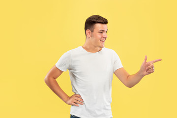 Caucasian young man's half-length portrait on yellow studio background. Beautiful male model in shirt. Concept of human emotions, facial expression, sales, ad. Pointing at side, looks happy.