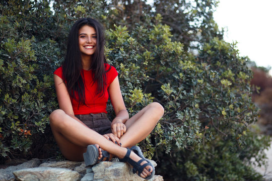 Beautiful girl, with long hair, wear in red, sitting on the rock, sitting near trees and rocks on the beach in Corsica.
