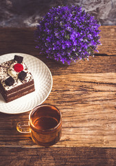 Hot tea In clear glass, black forest creek cake decorate with cherry and cake brownies In a white plate, and have false heather In pots small, all place are on wooden table.