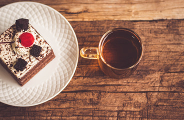 Hot tea In clear glass, black forest creek cake decorate with cherry and cake brownies In a white plate, and have false heather In pots small, all place are on wooden table.
