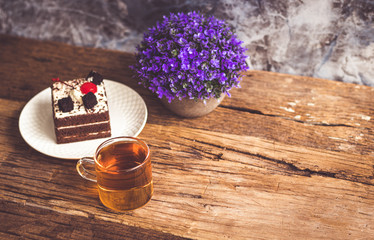 Hot tea In clear glass, black forest creek cake decorate with cherry and cake brownies In a white plate, and have false heather In pots small, all place are on wooden table.