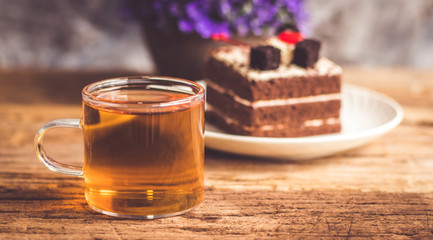 Hot tea In clear glass, black forest creek cake decorate with cherry and cake brownies In a white plate, and have false heather In pots small, all place are on wooden table.
