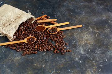 Roasted coffee beans spill out of the bag onto a dark textured surface. Wooden spoons with ground...