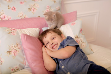 Obraz na płótnie Canvas A smiling happy boy lies on a bed with Shebby-chic bedding. British kittens frolic around him.