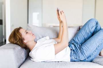 Young brunette woman in casualwear and headphones scrolling in smartphone while listening to music on couch