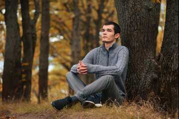 Obraz premium A young Man in a knitted sweater sitting in the autumn forest by a tree. Lifestyle autumn