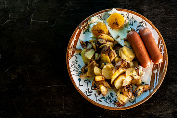 a plate with fried potatoes, scrambled eggs and sausages on dark background